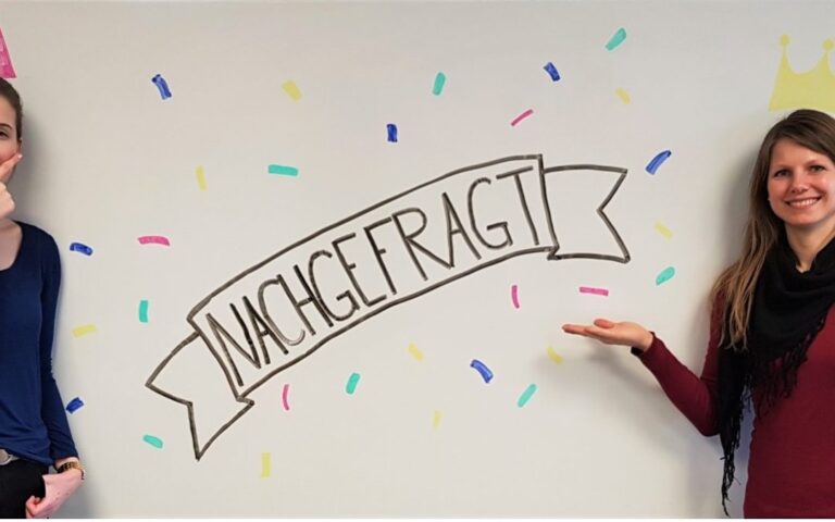 Wandbild „Nachgefragt“, zwei Mitarbeitende zeigen auf das Banner