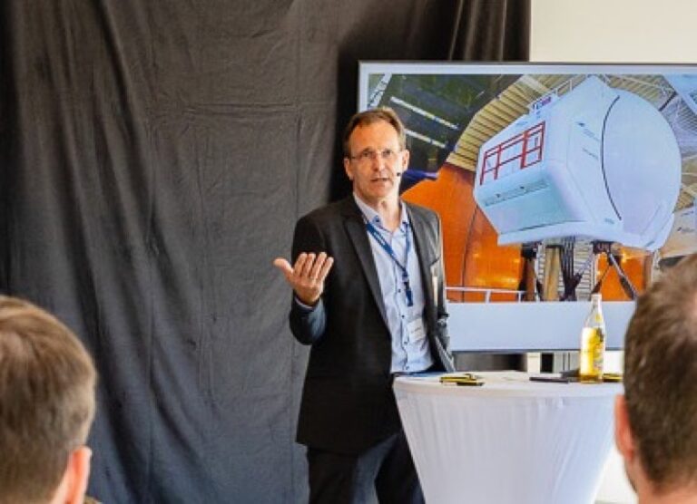 Jens Jährling von Lufthansa Aviation Training spricht vor Publikum, auf der Leinwand der H145-XR-Simulator von REISER